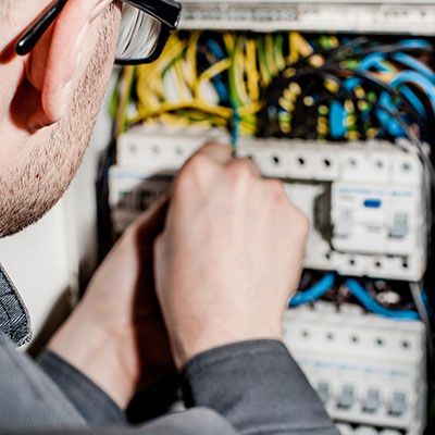 En person med kort hår och glasögon arbetar på en elektrisk panel och justerar kablar. Panelen är fylld med kablar och strömbrytare i olika färger. Personen har en mörk långärmad tröja på sig. Fokus ligger på deras händer och de intrikata ledningarna.